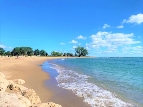 A serene beach scene with golden sand, gentle waves, and a clear blue sky dotted with clouds. Trees line the shore.