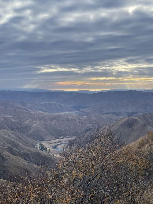 A panoramic view of rolling hills and valleys under a cloudy sky, with a river winding through the landscape at sunset.