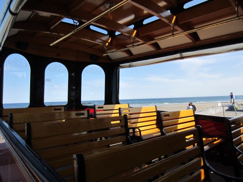 Interior of a trolley with wooden benches, overlooking a beach and ocean under a clear blue sky.