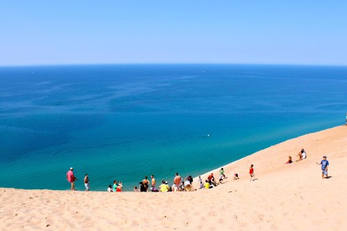 A sandy dune overlooking a blue lake, with a group of people enjoying the view and relaxing on the slope.