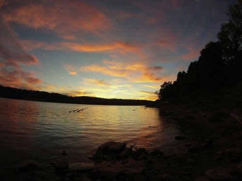 A serene lake at sunset, with colorful clouds reflecting on the water and a forested shoreline in the background.