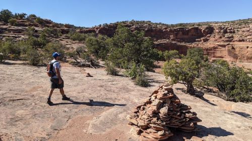 A person walks on rocky terrain near a stacked stone cairn, surrounded by shrubs and cliffs under a clear blue sky.