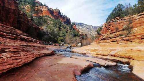 A serene river flows through red rock formations and lush greenery under a blue sky in a canyon landscape.