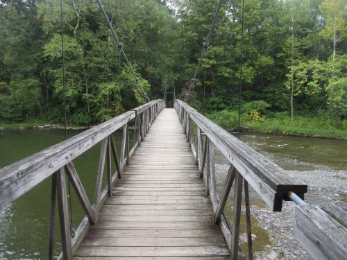 A wooden suspension bridge spans a calm river, surrounded by lush green trees and foliage.