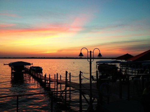 A serene sunset over a calm lake, with boats docked and silhouettes of a pier and lampposts against the colorful sky.