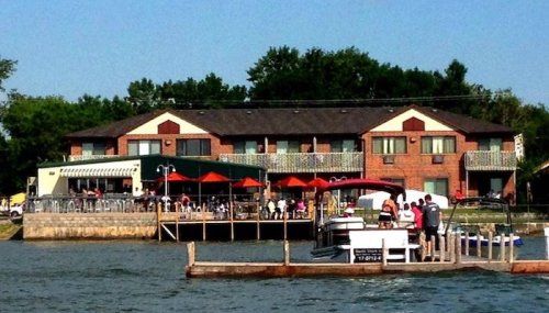 A waterfront restaurant with outdoor seating, surrounded by trees and a dock with boats on a sunny day.