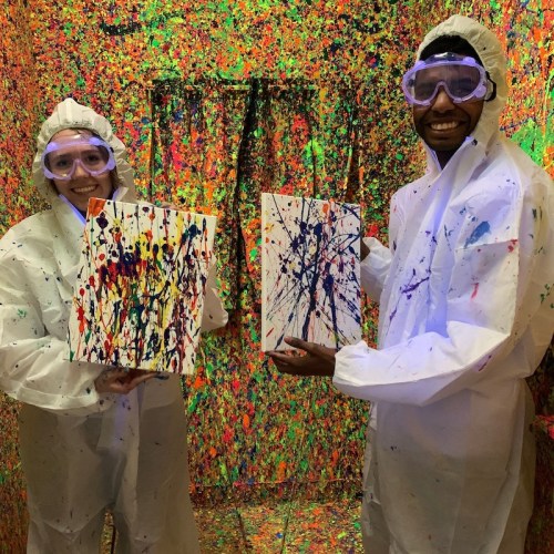 Two people in protective gear hold colorful splatter paintings in a vibrant, paint-splattered room.