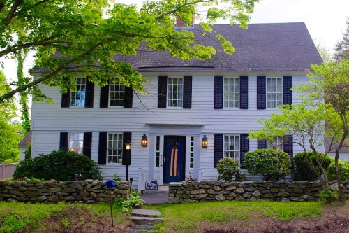 A charming white colonial-style house with black shutters, surrounded by greenery and a stone wall.