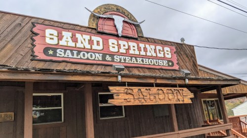 Sign for "Sand Springs Saloon & Steakhouse" on a rustic wooden building with a bull horn decoration.