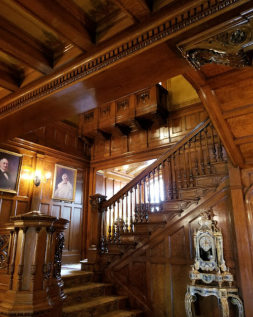 A grand wooden staircase with ornate details, framed portraits, and rich wood paneling in an elegant interior.