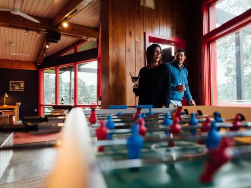 A couple enjoys drinks in a cozy room with large windows, standing near a foosball table.