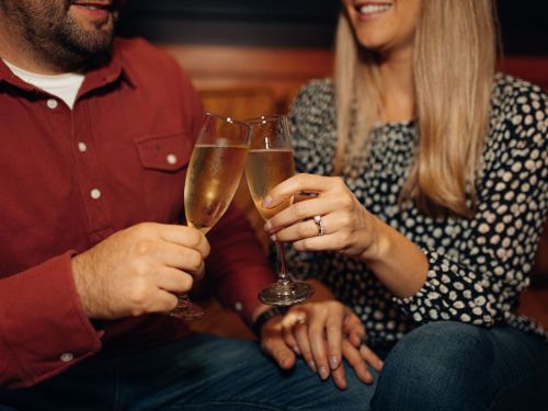 A couple clinks champagne glasses together, smiling, with one partner showing a ring on their hand.