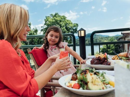 A woman and a girl enjoy a meal outdoors, with drinks and dishes on the table, against a scenic backdrop.