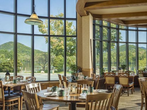 Bright dining area with wooden tables and chairs, large windows showcasing a scenic view of mountains and a lake.