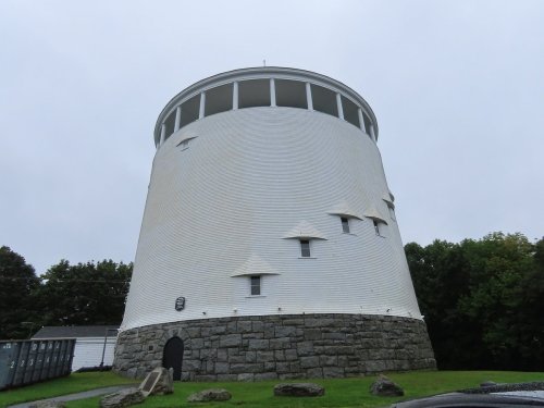 A tall, white cylindrical building with small windows, set against a cloudy sky and surrounded by greenery.