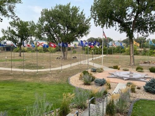 A park with a pathway lined by various flags and trees, featuring a landscaped area with plants and a circular stone design.