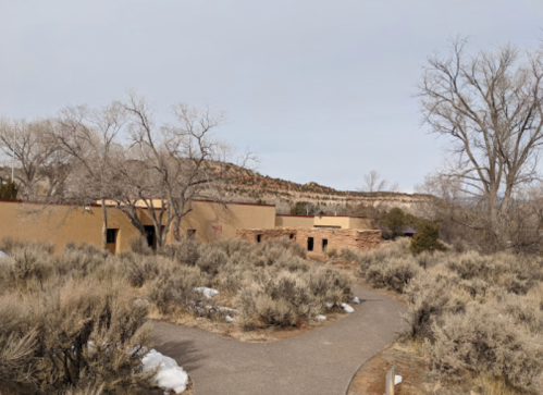 A winding path leads through sparse vegetation to adobe-style buildings against a backdrop of hills and bare trees.