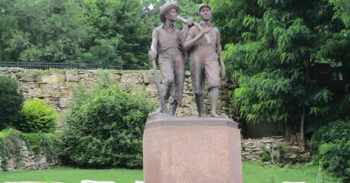 Statue of two boys walking together, one carrying a stick, set against a lush green background.