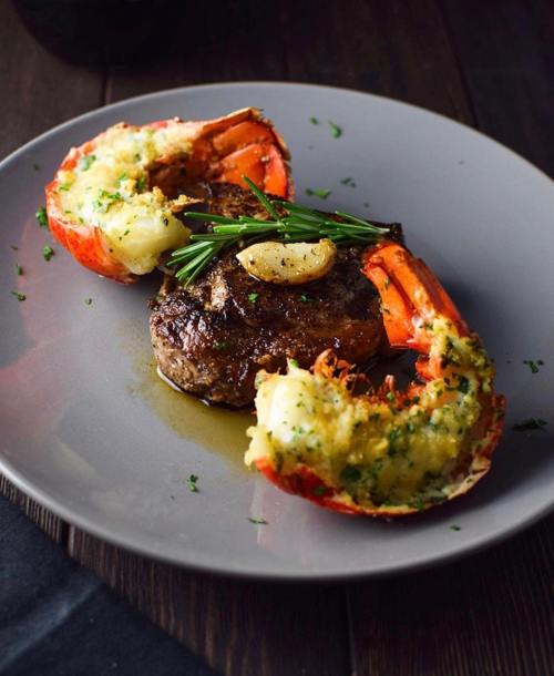 A plate featuring a grilled steak alongside two lobster tails, garnished with herbs and garlic.