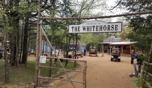 Entrance to The Whitehorse, featuring a rustic sign and a wooded area with picnic tables and a building in the background.