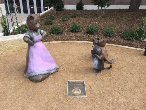 A bronze statue of a bear in a dress stands with two smaller raccoon figures in a landscaped area.
