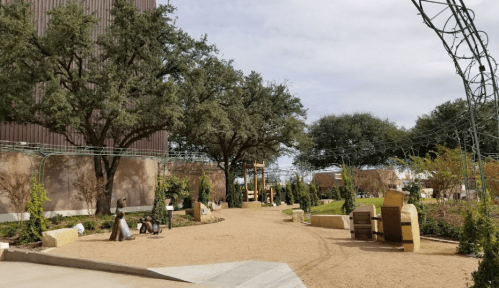A landscaped garden path with trees, sculptures, and seating areas under a cloudy sky.
