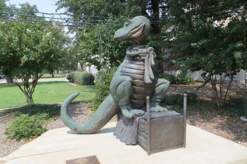 A bronze statue of a cartoonish dinosaur sitting on a box, surrounded by greenery.