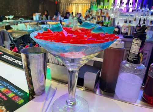 A colorful cocktail in a large glass, topped with red and blue jelly candies, sits on a bar counter.