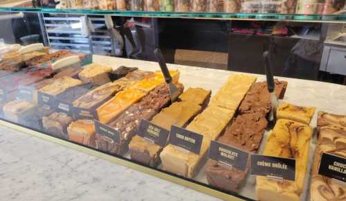 A display of various fudge flavors in a glass case, with labels and serving utensils.