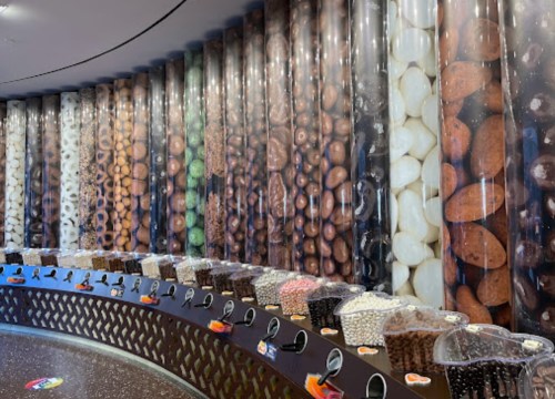 A colorful display of various candies and nuts in clear containers against a wall of vibrant, textured backgrounds.