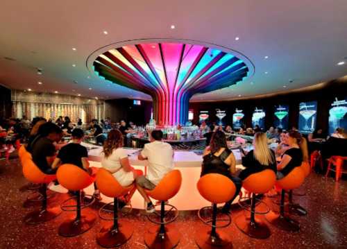 A vibrant bar scene with colorful lighting, featuring patrons seated on orange stools around a circular bar.