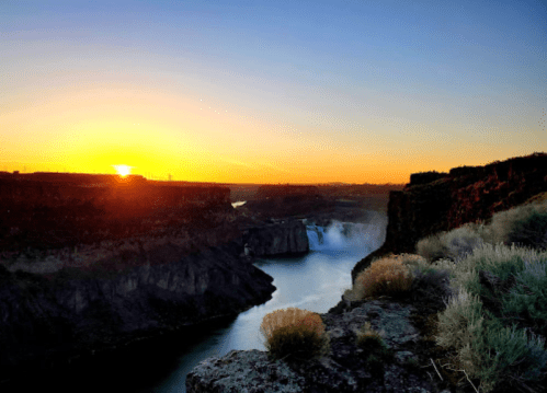 Sunrise over a canyon with a river, surrounded by rocky cliffs and sparse vegetation.