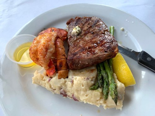 A plate featuring grilled steak, shrimp, asparagus, mashed potatoes, and a lemon wedge, with a small cup of sauce.