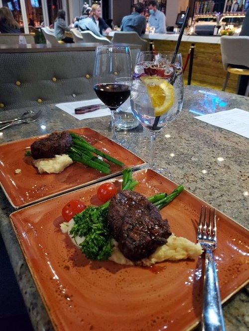 Two plates of steak with mashed potatoes, asparagus, and cherry tomatoes, accompanied by a glass of red wine and a cocktail.