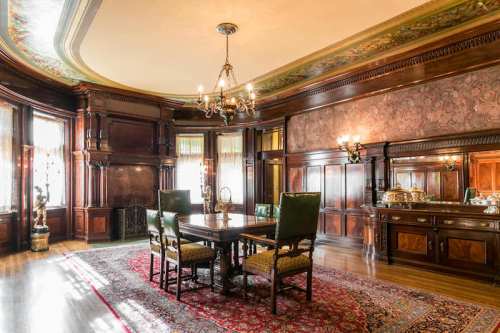 Elegant dining room with wooden paneling, a chandelier, and a large table surrounded by green chairs on a patterned rug.