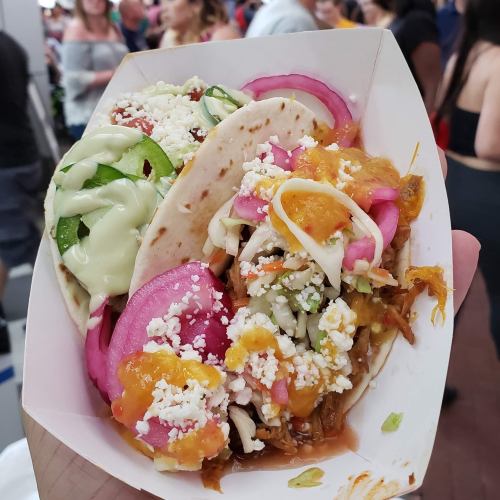 A hand holding a tray with two tacos topped with colorful ingredients like onions, cheese, and sauce, in a busy setting.