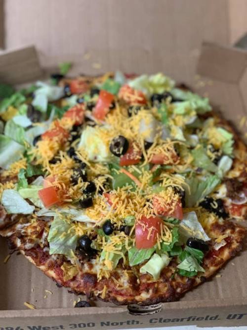 A pizza topped with lettuce, tomatoes, black olives, and shredded cheese, served in a pizza box.
