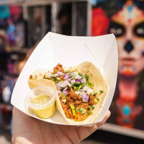 A hand holds a taco filled with meat, onions, and cilantro, alongside a small container of sauce, with a colorful mural in the background.