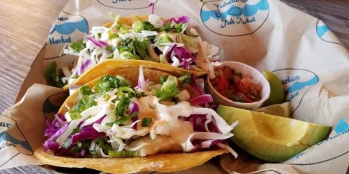 Two fish tacos topped with cabbage, cilantro, and sauce, served with lime wedges and a side of salsa in a basket.