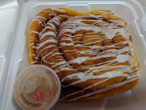 A cinnamon roll drizzled with icing, served in a takeout container with a small cup of icing on the side.