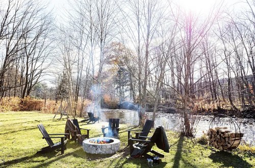 A cozy outdoor scene with a fire pit surrounded by chairs, near a river and bare trees under a bright sky.