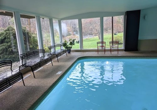 Indoor swimming pool with clear water, surrounded by large windows and seating areas, overlooking a green landscape.