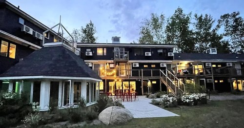 A cozy lodge at dusk, featuring multiple levels, outdoor seating, and surrounded by trees and gardens.