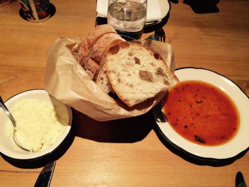 A basket of bread slices next to a bowl of tomato soup and a small dish of butter on a wooden table.