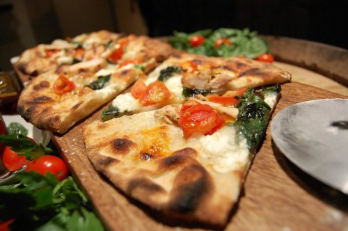A wooden platter with sliced pizza topped with spinach, tomatoes, and chicken, surrounded by fresh herbs and cherry tomatoes.