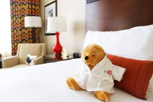A teddy bear in a white robe sits on a neatly made hotel bed, with colorful curtains and a lamp in the background.