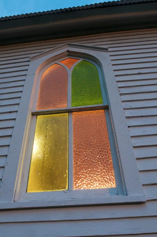 A stained glass window with colorful sections in green, orange, and yellow, set in a wooden building.