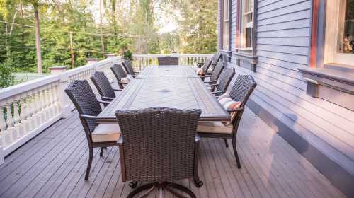 A spacious outdoor dining area with a long table and multiple chairs on a wooden deck surrounded by greenery.