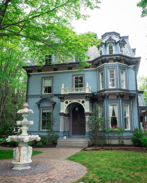 A charming blue Victorian house with intricate details, surrounded by greenery and featuring a decorative fountain in the front yard.