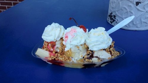 A colorful ice cream sundae with whipped cream, nuts, cherries, and a banana in a clear dish on a blue table.